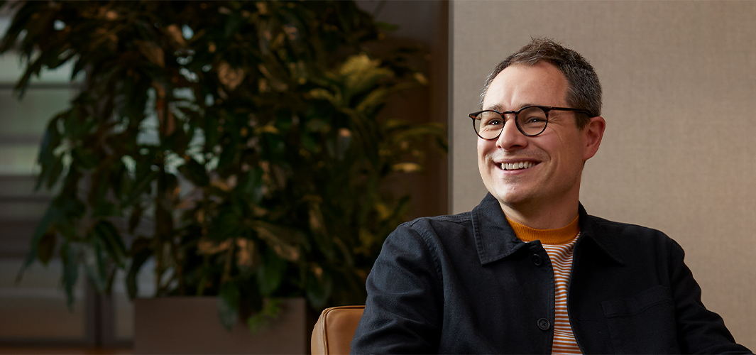 Dan lächelt, sitzt auf einem Sessel und sieht jemanden rechts neben sich an.