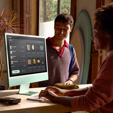 Un dipendente seduto dietro il bancone di un negozio che vende tavole da surf e altri articoli mentre usa un iMac per completare un acquisto per il cliente che ha di fronte.