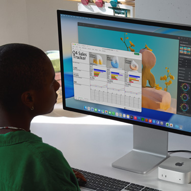 Employee sits and works on an Excel spreadsheet titled “Q4 Sales Tracker” on iMac and Mac mini. Behind the Excel spreadsheet is a Pixelmator Pro window and the macOS Tahoe background. 