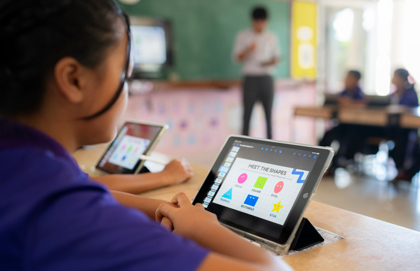 Close-up of a student at their desk, using an iPad. On screen, there are colourful shapes displayed with the title, “Meet the shapes”.”.