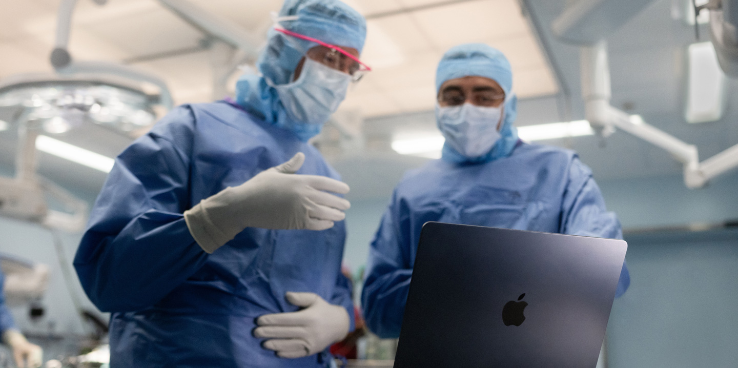 Deux chirurgiens sont dans une salle d’opération et regardent un MacBook ; l’un fait un geste de la main tandis que l’autre observe