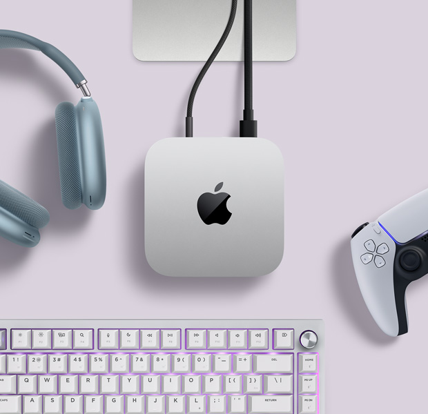Skats no augšas uz sudraba krāsas Mac mini, kura aizmugures portos iesprausti divi vadi un kuram līdzās novietotas zilas AirPods Max austiņas, sudraba Magic Keyboard tastatūra un bezvadu spēļu pults